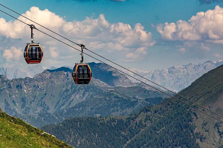 Zwei-rot-schwarze-Gondeln-der-Stubnerkogelbahn-schweben-vor-einer-alpinen-Bergkulisse-unter-leicht-bewoelktem-blauem-Himmel