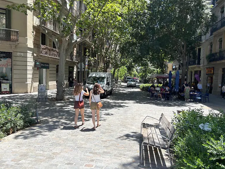 Zwei-Frauen-schlendern-die-schattige-Strasse-Girona-in-Barcelonas-Stadtteil-Eixample-entlang-Hier-in-einer-der-verkehrsberuhigten-Oasen-aus-mehreren-Haeuserbloecken-koennen-die-Menschen-gerade-auch-bei-Hitzewellen-im-Schatten-grosser-Baeume-ausruhen
