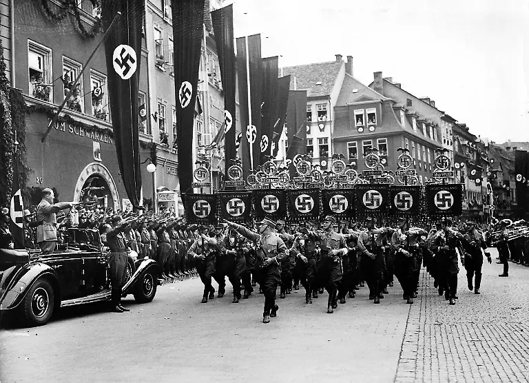 Adolf-Hitler-gruesst-die-Standarten-der-SA-Formationen-auf-dem-Erinnerungsparteitag-1936-der-zum-Gedenken-an-das-zehnjaehrige-Jubilaeum-des-ersten-Parteitags-nach-der-Neugruendung-der-NSDAP-in-Weimar-stattfand-1926-war-Hitler-in-Bayern-mit-einem-Redeverbot-belegt-deshalb-war-der-Parteitag-nach-Weimar-verlegt-worden-Alle-weiteren-fanden-dann-in-Nuernberg-statt-Vor-Hitlers-Auto-der-Stellertreter-des-Fuehrers-Reichsminister-Rudolf-Hess-Hinter-Hitler-die-sog-Blutfahne-der-Partei-Aufnahmedatum-01-07-1936-31-07