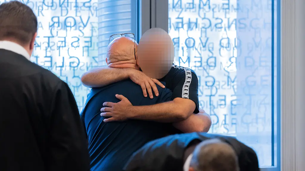 Der-Hauptangeklagte-l-und-ein-Angeklagter-umarmen-sich-in-einem-Gerichtssaal-im-Justizzentrum-Der-Prozess-um-ein-grosses-Waffenlager-von-Remscheid-ist-gestartet-Angeklagt-sind-ein-60-Jaehriger-sowie-zwei-Mitangeklagte-im-Alter-von-38-und-35-Jahren-Nach-Angaben-der-Staatsanwaltschaft-soll-der-60-Jaehrige-zahlreiche-Kriegswaffen-Schusswaffen-und-Munition-zum-Verkauf-gelagert-haben-Laut-Staatsanwaltschaft-handelt-es-sich-um-einen-der-groessten-Kriegswaffenfunde-der-vergangenen-Jahrzehnte-in-Deutschland