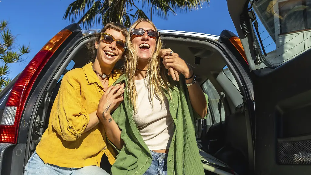 Zwei-laechelnde-Freunde-sitzen-mit-einem-Surfbrett-in-ihrem-Kofferraum-und-geniessen-einen-sonnigen-Sommer-Roadtrip-in-Australien-bereit-fuer-ein-unbeschwertes-Strandsurf-Abenteuer-zusammen-Modellfreigabe-vorhanden