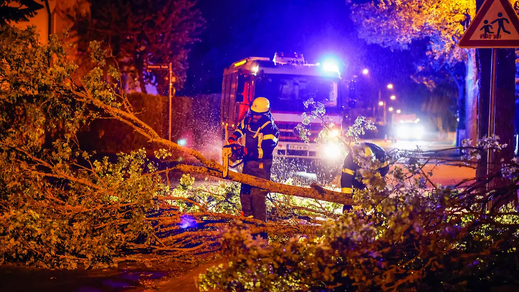 Einsatzkraefte-der-Feuerwehr-zersaegen-mit-einer-Kettensaege-einen-umgestuerzten-Baum-auf-einer-Strasse-im-Regierungsbezirk-Mittelfranken-Das-ueber-Deutschland-hinweggezogene-Unwetter-hat-am-Montagabend-in-Teilen-des-Landes-unter-anderem-Baeume-umstuerzen-lassen-und-Daecher-abgedeckt