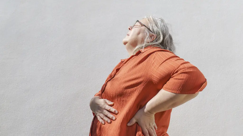 Aeltere-Frau-steht-draussen-an-einer-weissen-Wand-fuehlt-sich-unwohl-und-reibt-sich-den-unteren-Ruecken-um-das-Konzept-von-Schmerzen-Gesundheitsproblemen-und-Alterserscheinungen-zu-vermitteln-Modellfreigabe-vorhanden
