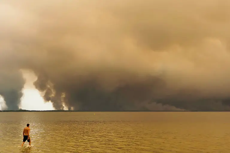 Ein-Strandbesucher-spaziert-am-Strand-von-Moutchic-in-Lacanau-im-Suedwesten-Frankreichs-am-Etang-de-Lacanau-entlang-waehrend-Rauchwolken-von-einem-Waldbrand-in-den-Himmel-aufsteigen-Das-Feuer-das-im-noerdlichen-Teil-des-Arcachon-Beckens-in-der-Gironde-im-Suedwesten-Frankreichs-wuetet-hat-nach-den-neuesten-Zahlen-der-Praefektur-bereits-mehr-als-3-400-Hektar-verwuestet