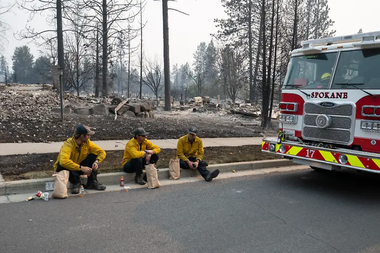 Three-members-of-the-Spokane-Fire-Department-sit-on-a-curb-in-front-of-burned-out-residential-homes-in-the-Balboa-neighborhood-of-Spokane-Washington-on-August-2-2026-Fast-moving-fires-left-a-trail-of-destruction-in-the-northwestern-US-city-of-Spokane-on-Sunday-with-officials-reporting-hundreds-of-structures-reduced-to-cinders-Thousands-of-people-were-ordered-to-evacuate-their-homes-Saturday-and-overnight-into-Sunday-as-three-separate-fires-broke-out-in-and-around-the-city-in-the-east-of-Washington-state