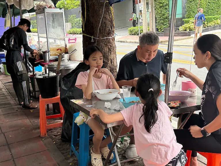 Eine-Familie-isst-an-einem-Strassenstand-in-Bangkok-Reisnudelsuppe-zum-Fruehstueck
