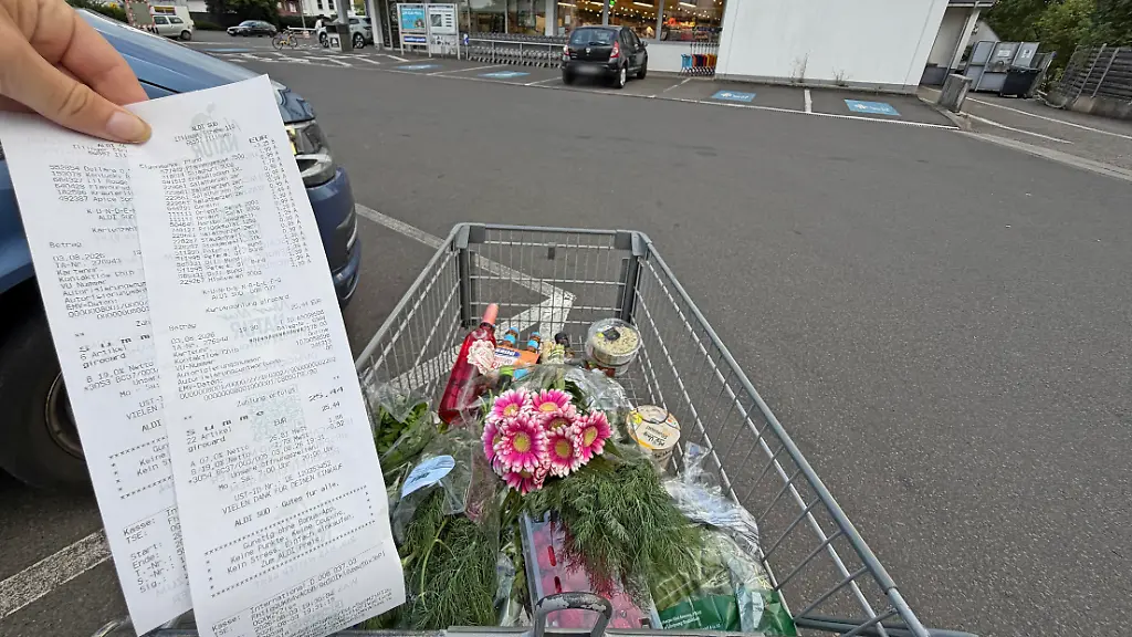 03-08-2026-Wirtschaft-v-l-Ein-gefuellter-Einkaufswagen-mit-Lebensmitteln-und-Blumen-steht-vor-einer-Aldi-Sued-Filiale-waehrend-ein-Kassenbon-in-die-Kamera-gehalten-wird-Das-Motiv-verdeutlicht-einen-typischen-Einkauf-im-Lebensmitteleinzelhandel-und-steht-fuer-Themen-wie-Einkaufen-Verbraucher-Preise-Lebensmittelversorgung-und-den-Alltag-im-Supermarkt