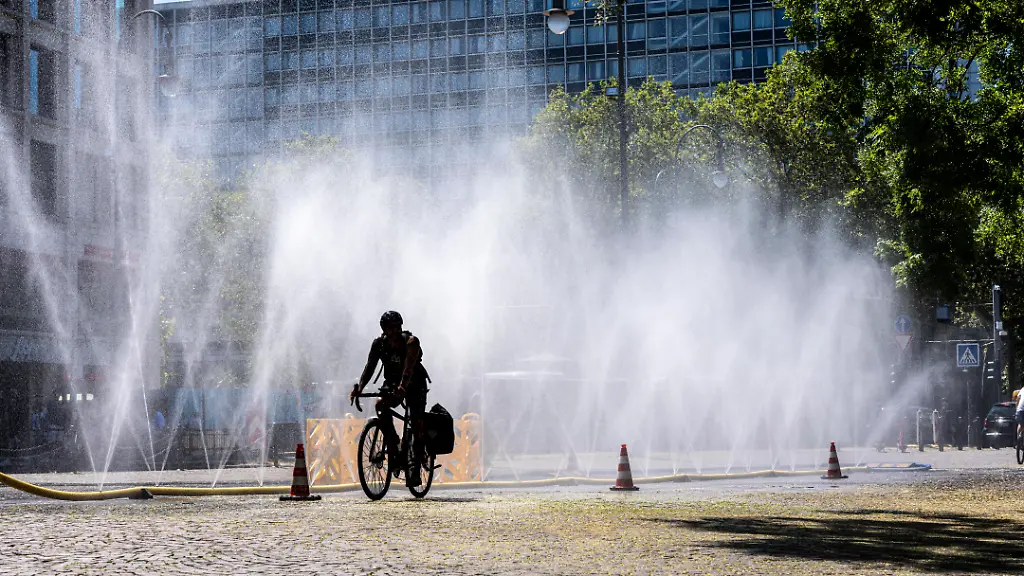 Cooling-Cologne-Aktion-der-Stadt-Koeln-zur-Abkuehlung-von-Hitzebelasteten-Plaetzen-hier-der-Rudolfplatz-in-der-Innenstadt-bei-knapp-37-C-an-dem-Tag-ein-Spruehschlauch-verspritzt-feine-Wassertroepfen-die-die-unmittelbare-Umgebung-einige-Grade-abkuehlt-eine-Aktion-die-bei-extremer-Hitzebelastung-vorgenommen-wird-zur-Anpassung-des-Stadtklimas-an-den-Klimawandel-Passanten-geniessen-den-kuehlenden-Moment-dazu-gibts-Tipps-zum-Umgang-mit-Extremhitze-NRW-Deutschland