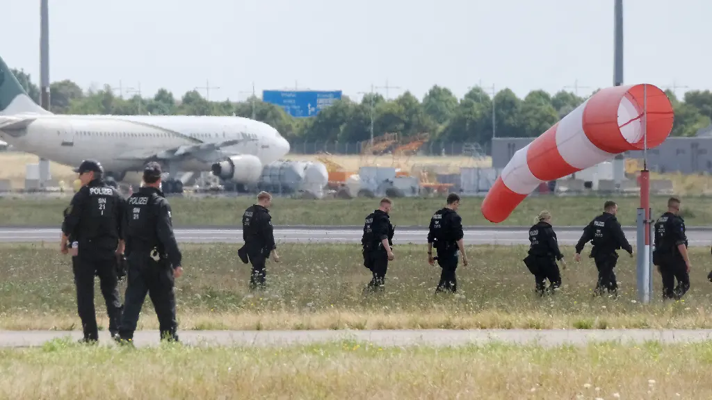 Polizisten-gehen-ueber-eine-Wiese-auf-dem-Gelaende-des-Flughafen-Leipzig-Die-Ermittlungen-nach-der-Entdeckung-der-Sprengstoff-Drohne-dauern-an-An-der-noerdlichen-Rollbahn-sind-zahlreiche-Polizistinnen-und-Polizisten-im-Einsatz-und-suchen-das-Gelaende-zu-Fuss-ab