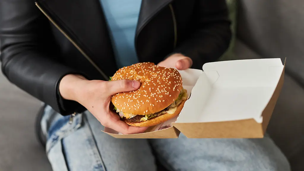 Close-up-shot-of-female-hands-holding-paper-box-with-delicious-cheeseburger-xkwx-woman-hand-tasty-burger-paper-box-eat-fast-food-delivery-meal-snack-lunch-delicious-appetizing-food-hamburger-meat-beef-cheese-vegetable-fresh-grill-junk-unhealthy-street-American-student-gen-Z-zoomer-teenager-adult-nutrition-leather-jacket-jeans-clothes-sit-couch-sofa-concept-unrecognizable-impersonal-home-indoors-background-copy-space