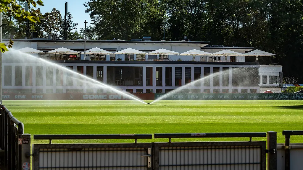Ein-Rasensprenger-laeuft-auf-dem-Trainingsgelaende-des-Fussball-Bundesligisten-1-FC-Koeln-Im-Hintergrund-ist-das-Geissbockheim-zu-sehen-Der-Ausbau-im-Gruenguertel-der-Stadt-ist-umstritten