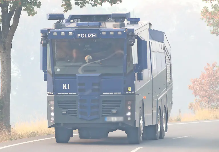 14-08-2026-Wirtschaft-v-l-Waldbrand-im-Huertgenwald-bei-Gey-Seit-dem-13-August-2026-kaempfen-zahlreiche-Einsatzkraefte-gegen-einen-grossflaechigen-Waldbrand-im-Huertgenwald-im-Kreis-Dueren-Nordrhein-Westfalen-Nach-Angaben-des-Kreises-Dueren-sind-ca-300-Hektar-Waldflaeche-betroffen-Etwa-400-500-Einsatzkraefte-verschiedener-Organisationen-sind-vor-Ort-Der-Ortsteil-Gey-wurde-aufgrund-der-Gefahrenlage-evakuiert-Die-Loescharbeiten-werden-unter-anderem-durch-schwieriges-Gelaende-und-moegliche-Munitionsaltlasten-aus-dem-Zweiten-Weltkrieg-erschwert-Die-Aufnahme-zeigt-Impressionen-aus-dem-Bereich-des-Waldbrandes-und-der-laufenden-Einsatzmassnahmen-am-14-August-2026-Auf-diesem-Foto-ist-ein-Wasserwerfer-der-Polizei-waehrend-des-Grosseinsatzes-rund-um-den-Waldbrand-im-Huertgenwald-zu-sehen-Im-Hintergrund-ist-die-starke-Rauchentwicklung-deutlich-erkennbar