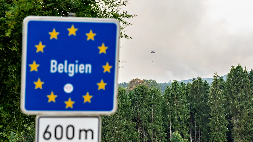 Blick-von-Kalterherberg-Gemeinde-Monschau-auf-den-Grossbrand-im-Hohen-Venn-nahe-der-deutsch-belgischen-Grenze-Im-Vordergrund-ein-blaues-Grenzschild-im-Hintergrund-Rauchsaeulen-des-Feuers-und-ein-Loeschhubschrauber-mit-Wasserbehaelter-im-Anflug-Insgesamt-zwei-Loeschhubschrauber-sind-im-Einsatz-und-werfen-jeweils-rund-3-000-Liter-Wasser-ab-Der-Hochmoorbrand-hat-bereits-rund-2-000-Hektar-Wald-und-Moorgebiet-erfasst-die-tiefen-Torfschichten-beguenstigen-einen-langanhaltenden-Schwelbrand-Auf-belgischer-Seite-laufen-bereits-Evakuierungen-fuer-Teile-Monschaus-wird-eine-moegliche-Raeumung-vorbereitet
