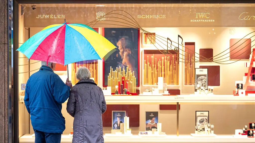Zwei-aeltere-Personen-stehen-bei-Regen-vor-einem-Schaufenster-eines-Schmuckgeschaefts-und-betrachten-die-ausgestellten-Waren-Sie-teilen-sich-einen-bunten-Regenschirm-Im-Bundestag-steht-am-Freitag-die-Entscheidung-ueber-die-geplante-Rentenreform-an-die-ein-stabiles-Rentenniveau-erweiterte-Kindererziehungszeiten-steuerfreie-Zuverdienstmoeglichkeiten-im-Alter-und-weitere-Aenderungen-in-der-Altersvorsorge-vorsieht