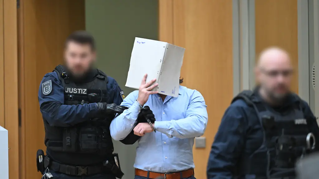 Einer-der-Angeklagten-M-in-einem-Staatsschutzverfahren-gegen-drei-Ukrainer-wegen-Agententaetigkeit-im-Auftrag-eines-russischen-Nachrichtendienstes-wird-von-Justizbeamten-in-den-Saal-des-Oberlandesgerichts-Stuttgart-in-Stammheim-gefuehrt-Die-ukrainischen-Angeklagten-sollen-im-Maerz-2025-ueber-einen-ukrainischen-Postdienstleister-zwei-Pakete-mit-GPS-Trackern-in-Richtung-Ukraine-versandt-haben-um-Versandrouten-und-Transportablaeufe-bei-dem-Paketdienstleister-auszuforschen
