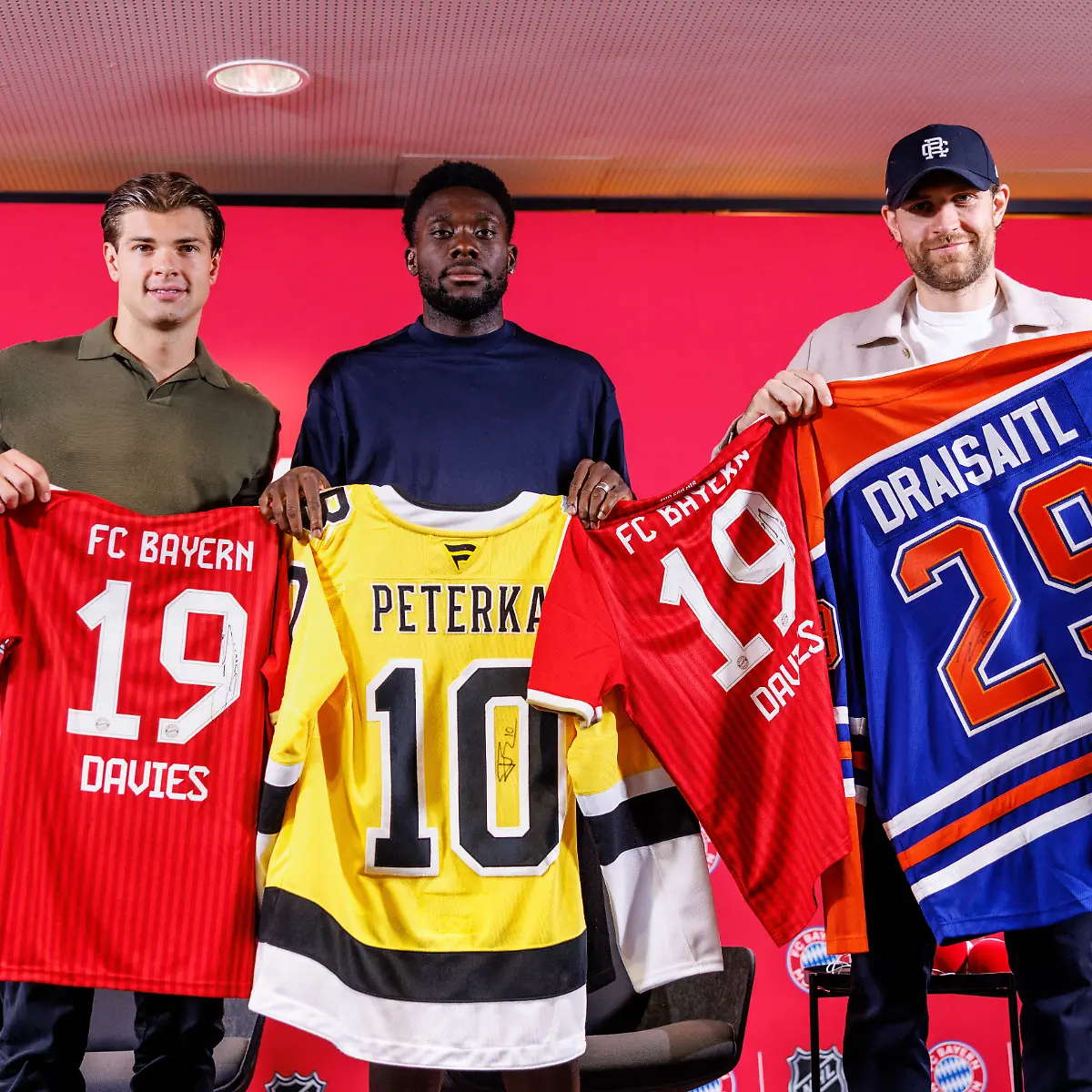 Eishockey-Fussball-Gemeinsame-Pk-des-FC-Bayern-Muenchen-und-der-National-Hockey-League-NHL-John-Jason-Peterka-l-r-Spieler-der-Boston-Bruins-Alphonso-Davies-Spieler-vom-FC-Bayern-Muenchen-und-Leon-Draisaitl-Spieler-der-Edmonton-Oilers-posieren-auf-der-Pressekonferenz-mit-Trikots