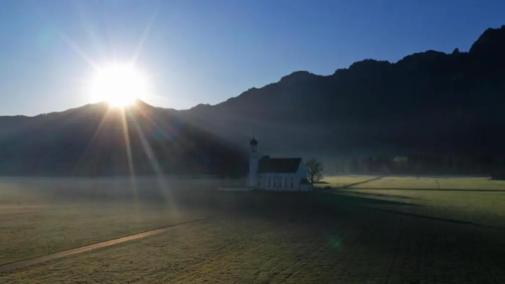 Die-Sonne-geht-ueber-den-Bergen-hinter-der-im-leichten-Morgennebel-liegenden-Wallfahrtskirche-Sankt-Coloman-auf