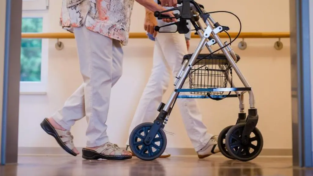 Eine-Pflegekraft-geht-in-einem-Pflegeheim-mit-einer-aelteren-Dame-ueber-einen-Korridor