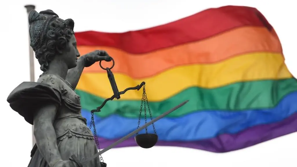 Eine-Regenbogenfahne-weht-hinter-der-Bronzestatue-der-roemischen-Goettin-der-Gerechtigkeit-Justitia-die-mit-einer-Waage-und-einem-Richtschwert-in-der-Hand-auf-dem-Gerechtigkeitsbrunnen-des-Roemerbergs-steht