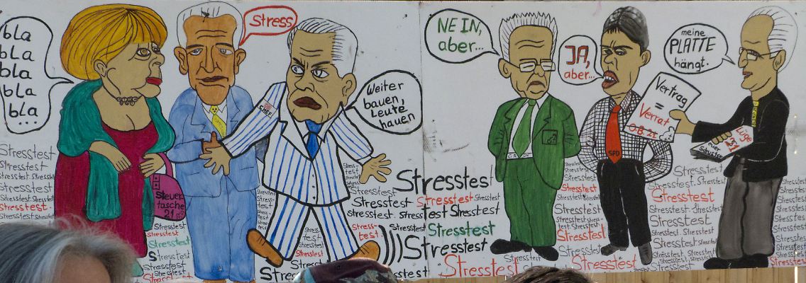 Teilnehmer der Montagsdemo hören den Reden zu, während hinter ihnen eine Karikatur zum Stresstest hängt.