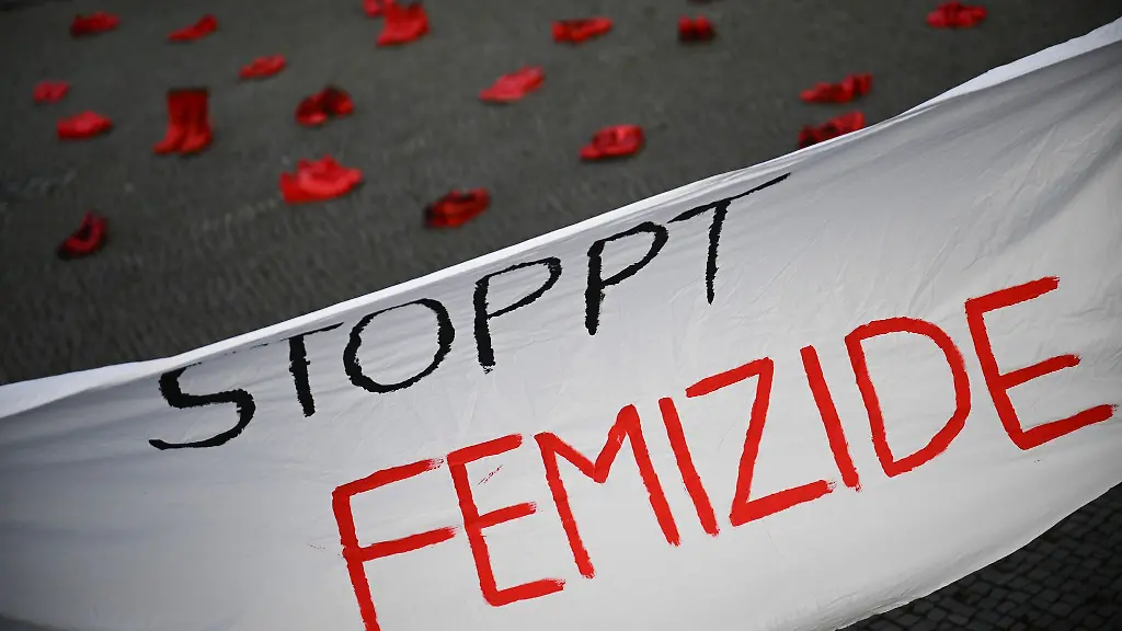 Ein-Banner-mit-der-Aufschrift-Stoppt-Femizide-haengt-vor-roten-Schuhen-zum-Internationalen-Tags-fuer-die-Beseitigung-von-Gewalt-gegen-Frauen