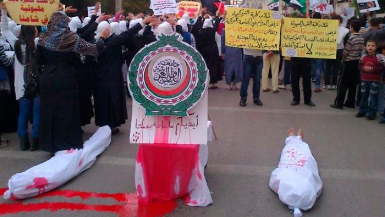 Proteste in Homs: Zwei Leichensäcke liegen neben einem Logo der Arabischen Liga.
