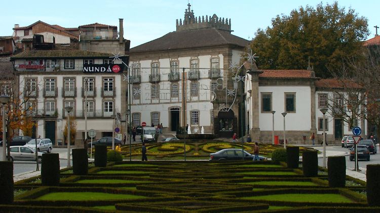 Blick über die Praca do Brasil, einen länglichen Platz im Zentrum von Guimarães. Im Hintergrund überragt der mit Zinnen bewehrte Glockenturm der Kirche Nossa Senhora da Oliveira die übrigen Bauten ringsum.
