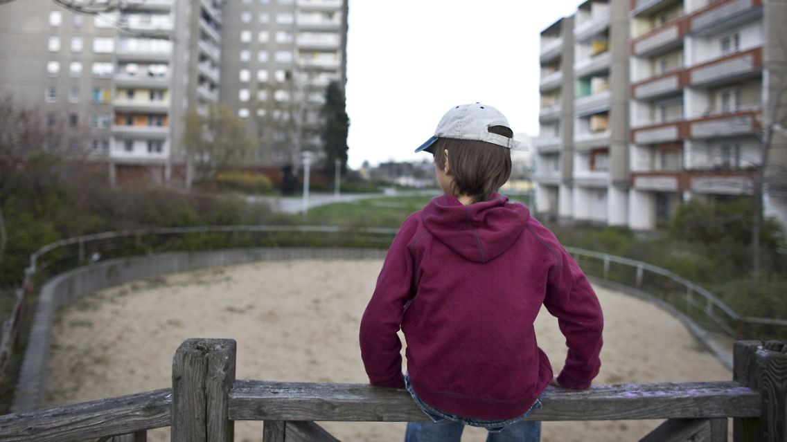 Ein kleiner Junge sitzt allein auf einem Spielplatzes im Innenhof eines Plattenbau-Wohngebiets in Frankfurt (Oder).