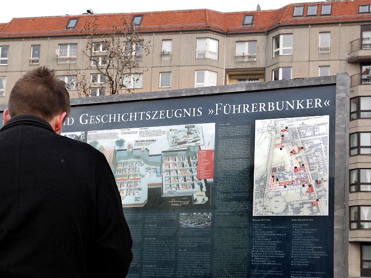 Tourist auf dem Gelände der ehemaligen Reichskanzlei und des Führerbunkers vor einer Informationstafel über die Geschichte des Areals. Im Hintergrund Wohnhäuser an der Wilhelmstraße.