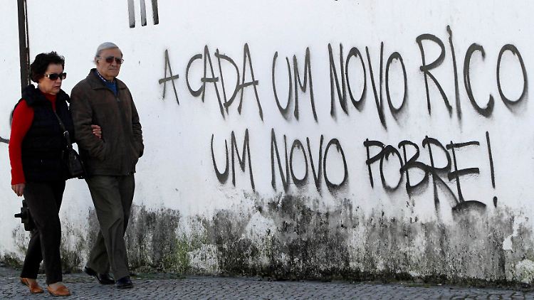 Ein Paar in Portugal läuft an einer Mauer mit dem Schriftzug "Für jeden neuen Reichen, ein neuer Armer" entlang.