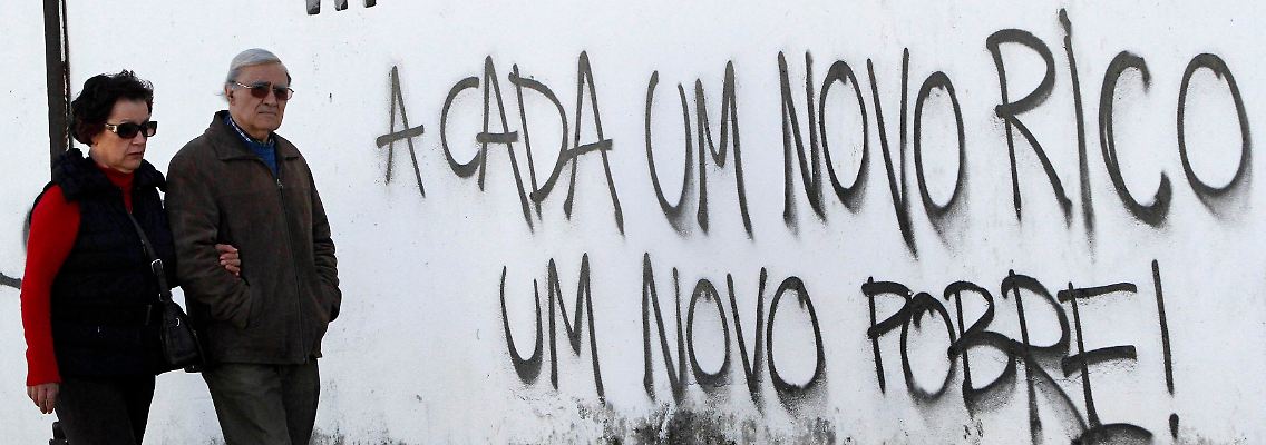 Ein Paar in Portugal läuft an einer Mauer mit dem Schriftzug "Für jeden neuen Reichen, ein neuer Armer" entlang.