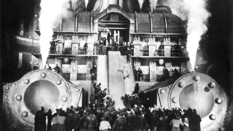 Eine aufgebrachte Menschenmenge steht vor einem dampfenden Maschinengebäude, eine Szene aus dem Stummfilmklassiger "Metropolis".