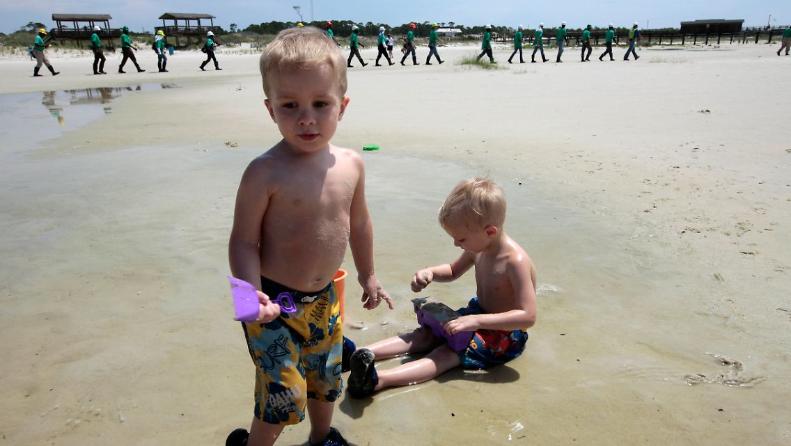 Kinder spielen am Strand von Dauphin Island, während im Hintergrund Reiningskräfte nach Ölspuren suchen.