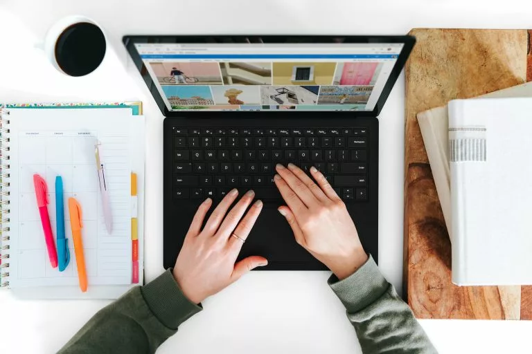 Blick von oben auf einen Schreibtisch auf dem ein aufgeklappter Laptop steht. Eine Person arbeitet daran. Neben dem Laptop liegt ein Block mit Stiften sowie Bücher. Auf deiner Hochzeits-Homepage dürfen ein paar Informationen nicht fehlen â Wir zeigen dir, welche das sind!