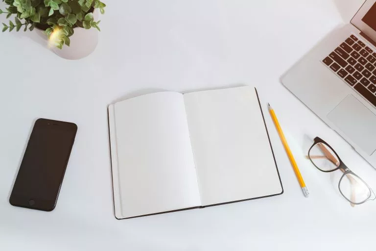 Auf einem weiÃen Schreibtisch liegt in der Bildmitte ein aufgeschlagenes Notizbuch mit leeren Seiten. Daneben liegt ein Handy, ein Bleistift und eine Brille. Wir zeigen dir, welche Anbieter sich für deine Hochzeits-Homepage lohnen!