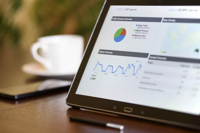 Nahaufnahme eines Tablets mit Diagrammen auf dem Bildschirm auf einem Tisch neben einem Handy und einer Kaffeetasse. Wir zeigen dir, wie du deine Hotel Website bekannter machst!