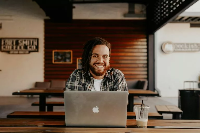 Ein junger Mann sitzt in einem hellen Raum vor einem Laptop an einem Tisch und lächelt in die Kamera. Mit unseren Tipps kannst du nicht nur eine Hotel-Website erstellen, sondern diese auch optimieren!