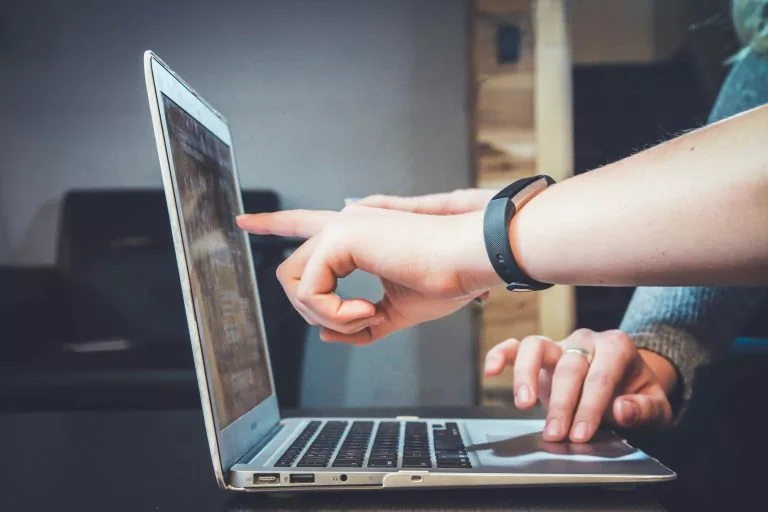 Blick von der Seite auf einen aufgeklappten Laptop. Eine Person bedient das Trackpad und eine andere Person zeigt auf den Bildschirm. Wenn du eine Portfolio Website erstellen willst, solltest du wichtige Entscheidungen zu Beginn treffen.
