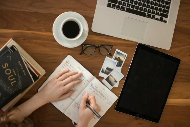 Blick von oben auf einen Schreibtisch. Eine Person schreibt etwas in ein Notizbuch. Daneben liegen Bücher, eine Brille sowie kleine Fotos. Am oberen Bildrand steht eine Tasse Kaffee neben einem aufgeklappten Laptop. Wir zeigen dir, worauf es beim Website Baukasten Vergleich ankommt!