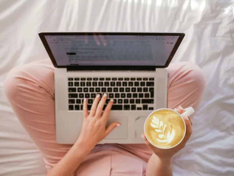 Blick von oben auf eine Frau, die in einem Bett sitzt und einen Laptop auf dem SchoÃ hat. In der rechten Hand hÃ¤lt sie eine Tasse Kaffee. Mit unserer Schritt-fÃ¼r-Schritt-Anleitung kannst du einfach einen Blog erstellen!