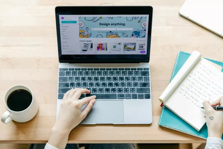 Blick von oben auf einen Holztisch auf dem ein aufgeklappter Laptop neben einer Kaffeetasse und einem Notizblock steht. Eine Person schreibt mit der rechten Hand in das Notizheft und tippt mit der linken Hand auf dem Trackpad vom Laptop. Wir zeigen dir, wie du ganz einfach eine perfekte Landingpage erstellen kannst!