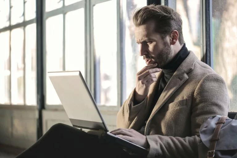 Blick auf einen sitzenden Mann im Anzug vor einer groÃÂen Fensterfront, der auf einen Laptop schaut und sich fragend den Bart streicht. Wir zeigen dir, ob ein Onepager zu dir passt!