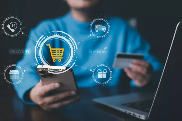 Eine Person in einem hellblauen Pullover sitzt in einem dunklen Raum vor einem aufgeklappten Laptop. Die Person hÃÂ¤lt in der rechten Hand ein Handy und in der linken Hand eine Bankkarte. Wir zeigen dir, wie du einen Onlineshop kostenlos erstellen kannst!