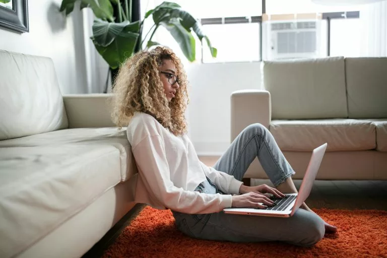 Eine junge Frau sitzt in einem hellen Raum auf dem Boden vor einem weiÃÂen Sofa mit einem Laptop auf dem SchoÃÂ. Wir zeigen dir, welche Anbieter sich lohnen, wenn du einen Onlineshop kostenlos erstellen willst!