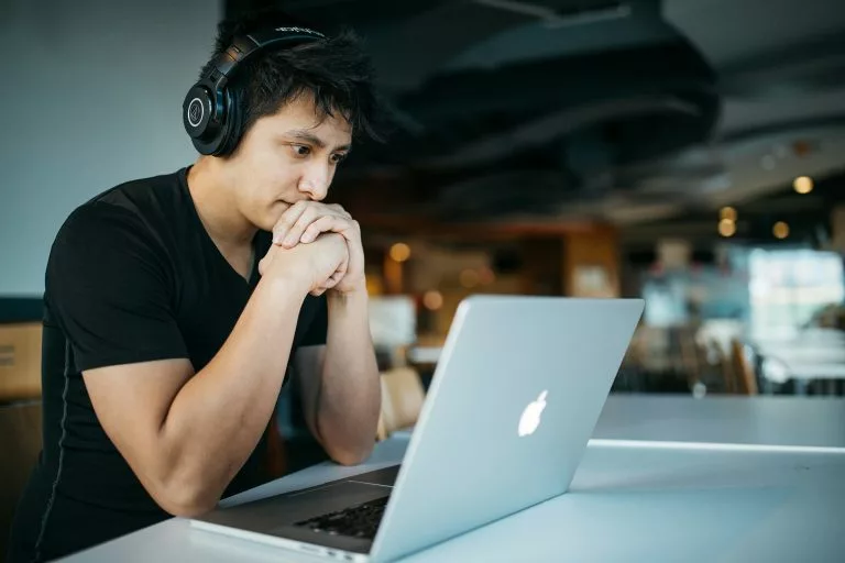Ein junger Mann mit Kopfhörern sitzt nachdenklich vor einem Laptop in einem GroÃÂraumbüro. Erfahre, wann es sich lohnt, einen Onlineshop kostenlos zu erstellen!