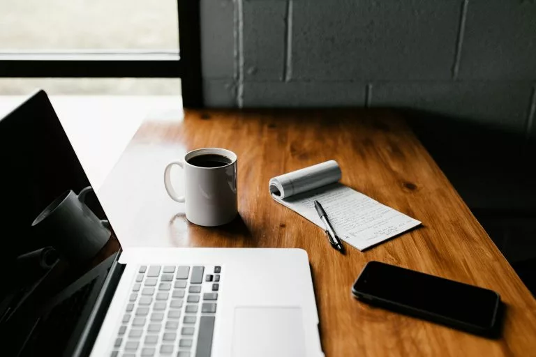 Auf einem Tisch stehen ein Laptop, Kaffee, Telefon und ein Zettel.